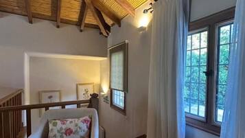Una habitación con techo de madera, una ventana con cortinas, una silla con cojín y un cuadro en la pared.