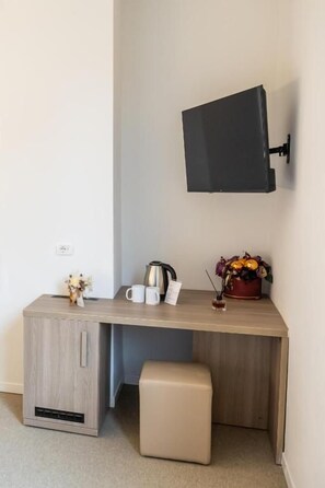 A small, compact room with a kitchenette area, a flat-screen TV mounted on the wall, and a small table with a coffee pot and flowers.