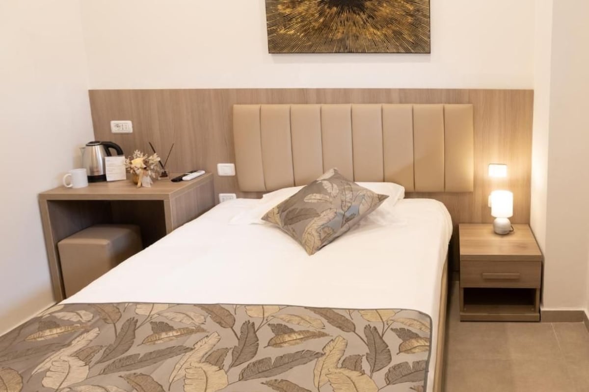 A hotel room with a bed, bedside tables, a nightstand, a lamp, and a wall-mounted artwork.