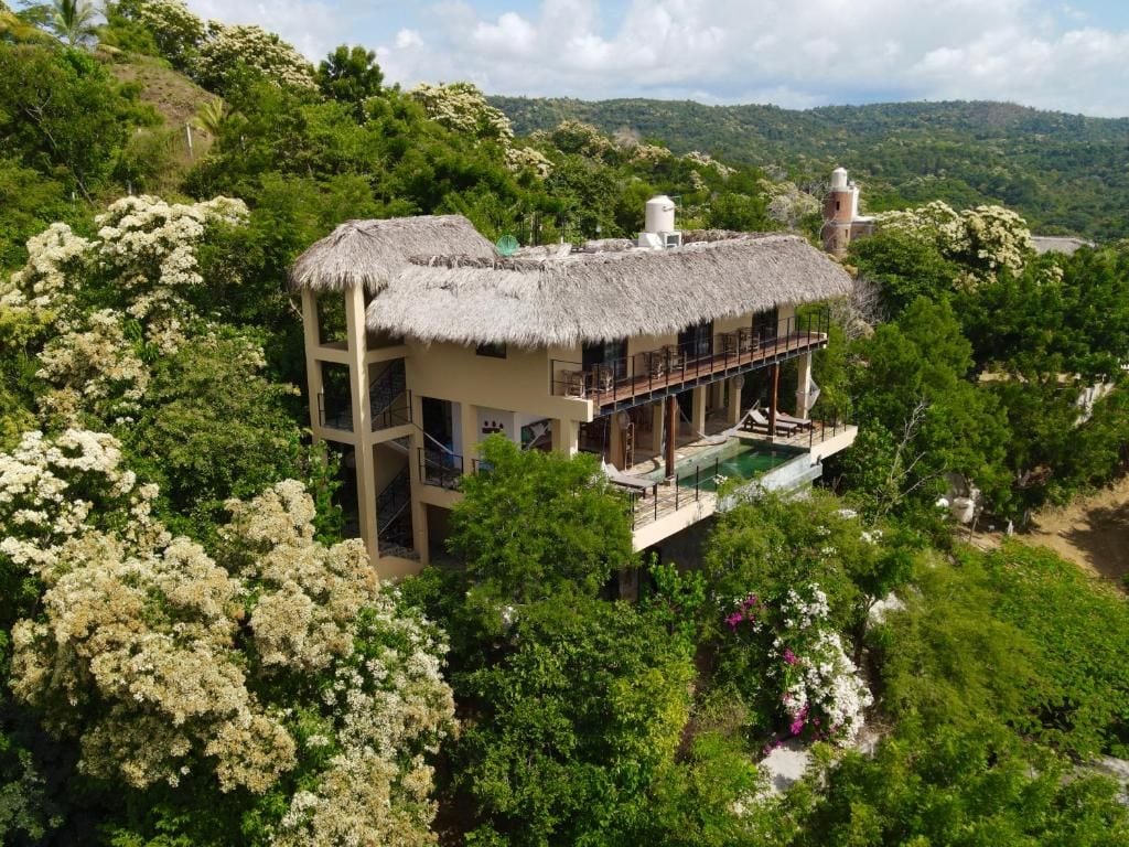 A thatched-roof house with a pool, surrounded by lush greenery and hills.