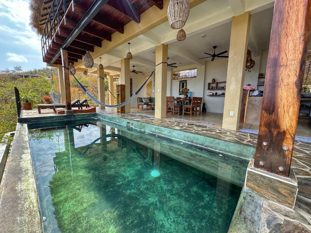 Una piscina cristalina con borde de piedra, un área de estar techada con sillas y mesas, y un ventilador de techo.