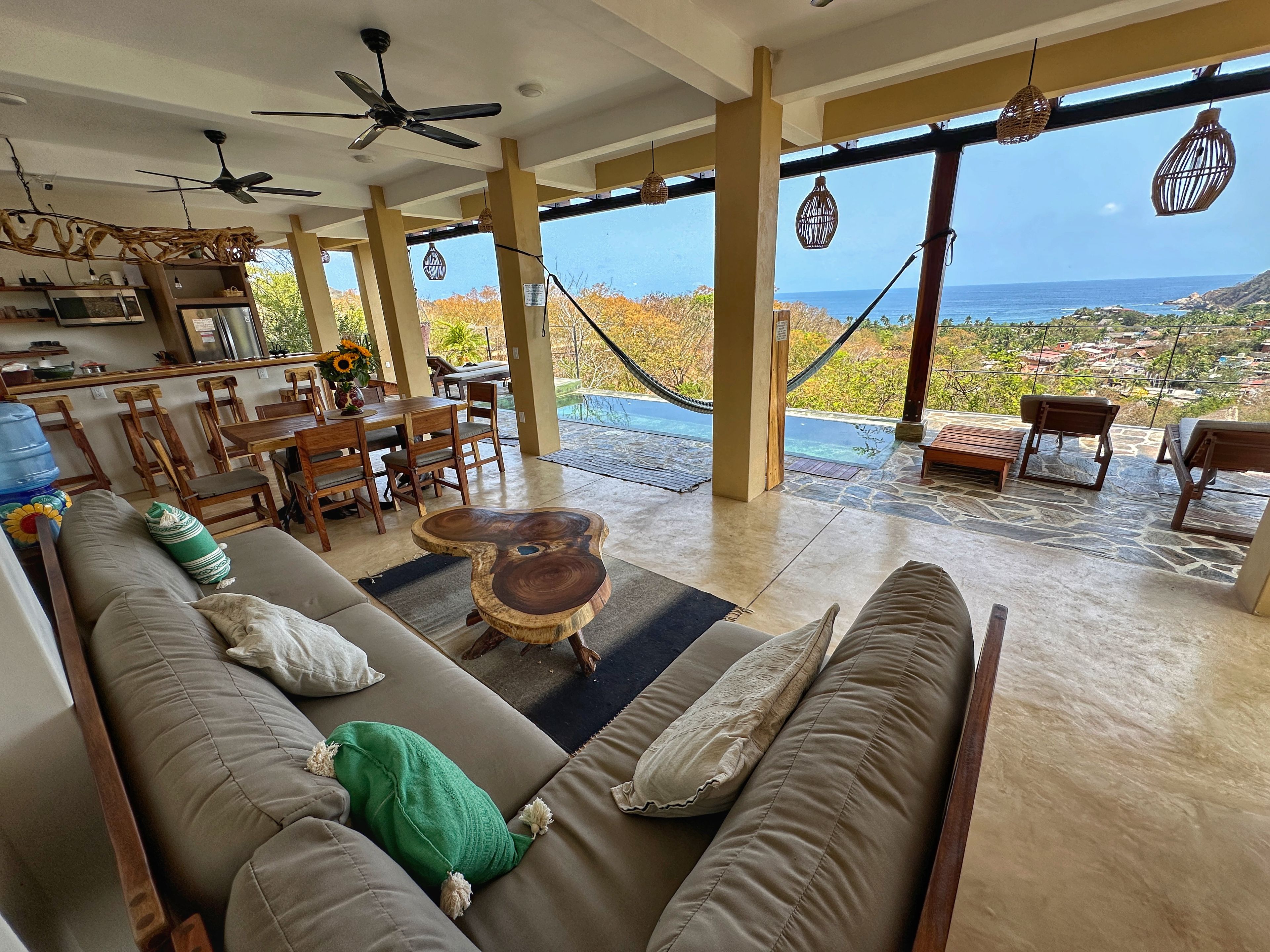 A spacious living area with a large sofa, dining table, and chairs, a wooden coffee table, and a view of the ocean through large windows.