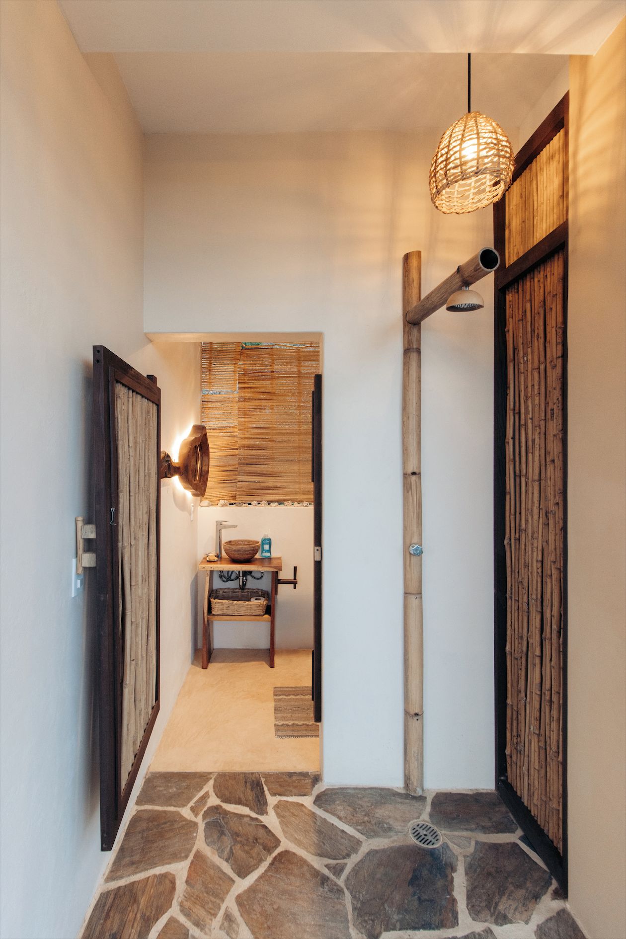 A hallway with a stone floor, wooden doors, a wall-mounted light fixture, and a small table with a basket and a bottle.