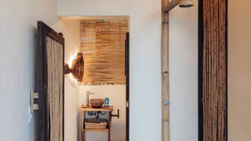 A hallway with a stone floor, wooden doors, a wall-mounted light fixture, and a small table with a basket and a bottle.