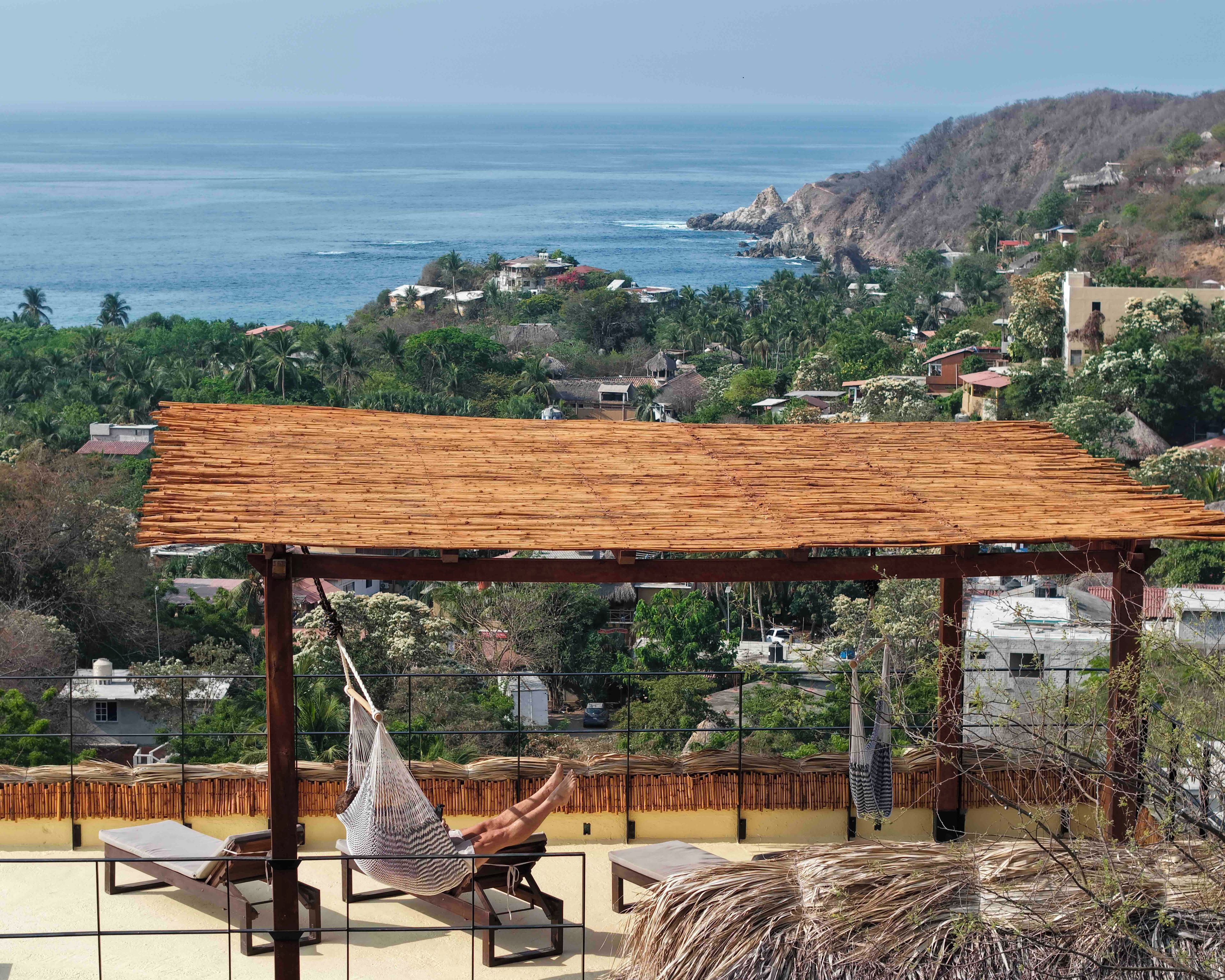 Eine Hängematte unter einer Holzpergola mit Blick auf eine Küstenstadt, mit Häusern und Palmen.
