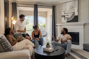 A group of people sitting on a couch and chairs in a well-lit living room with a fireplace, a large window, and a view of the outdoors. - Home Sweet Stay 2BD/2BA • Pool • Gym (Scottsdale)