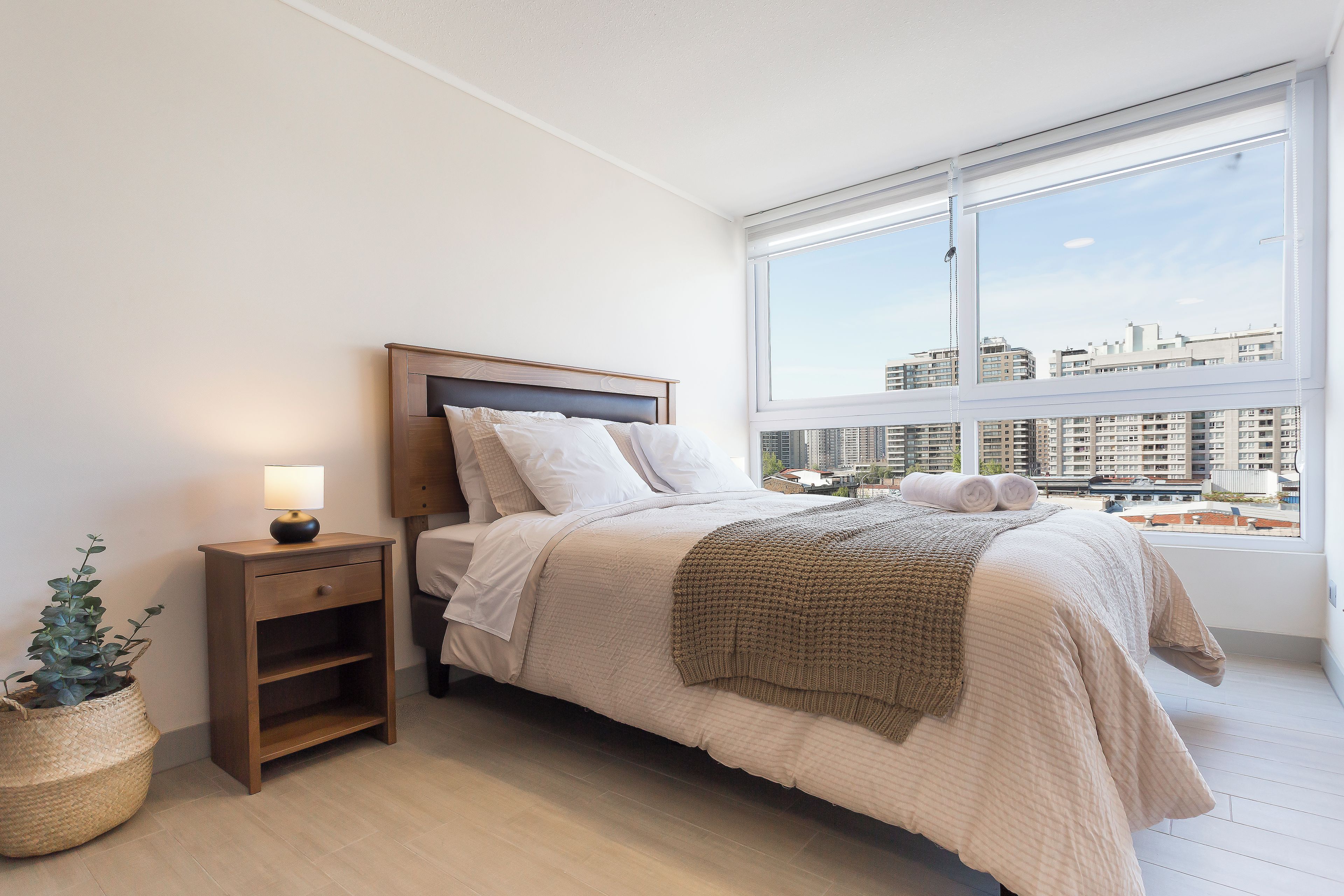 A bedroom with a bed, bedside table, lamp, and a view of the city through a large window.