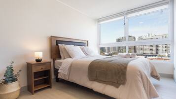 A bedroom with a bed, bedside table, lamp, and a view of the city through a large window.