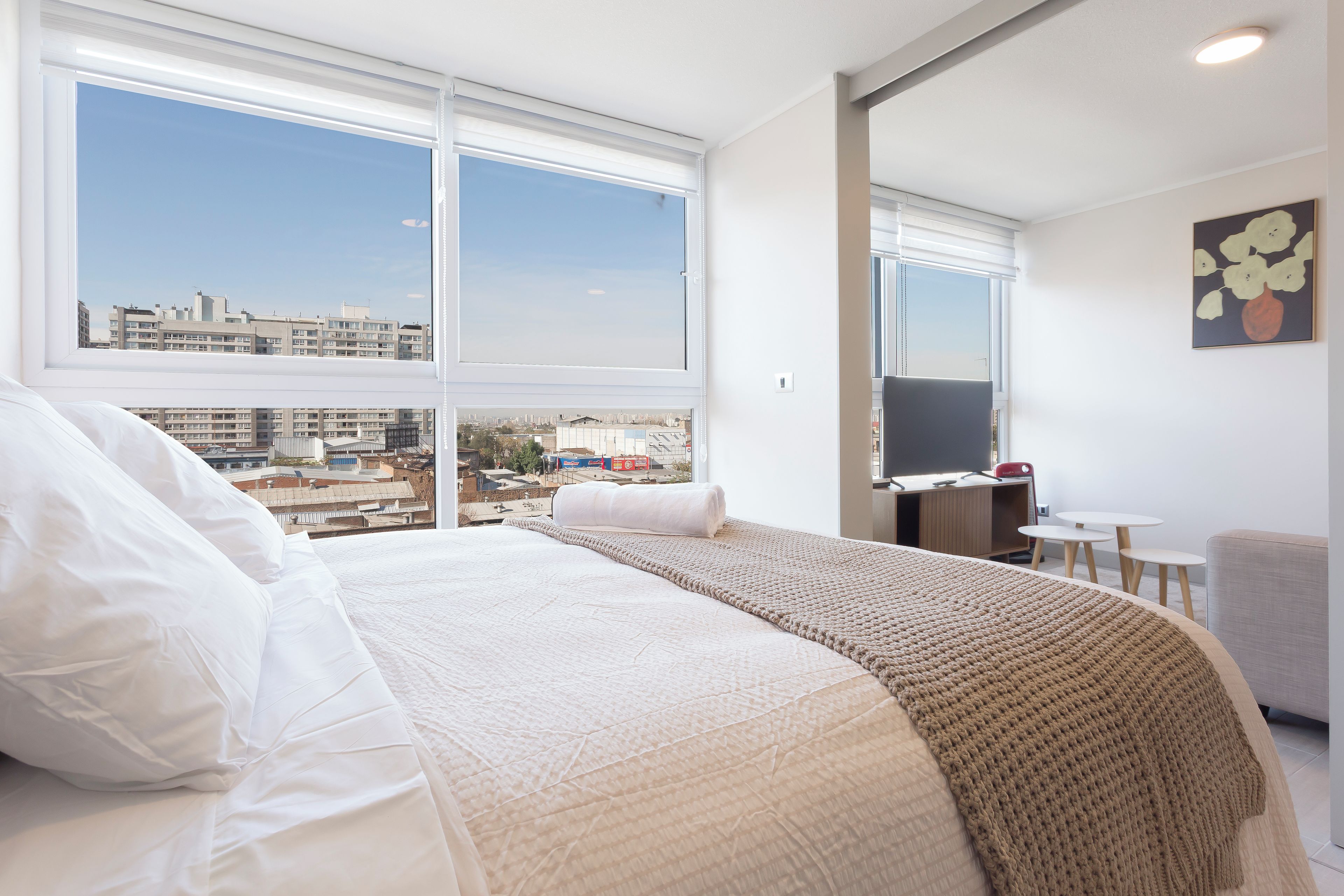 A hotel room with a large bed, a desk, a chair, and a view of the cityscape through large windows.