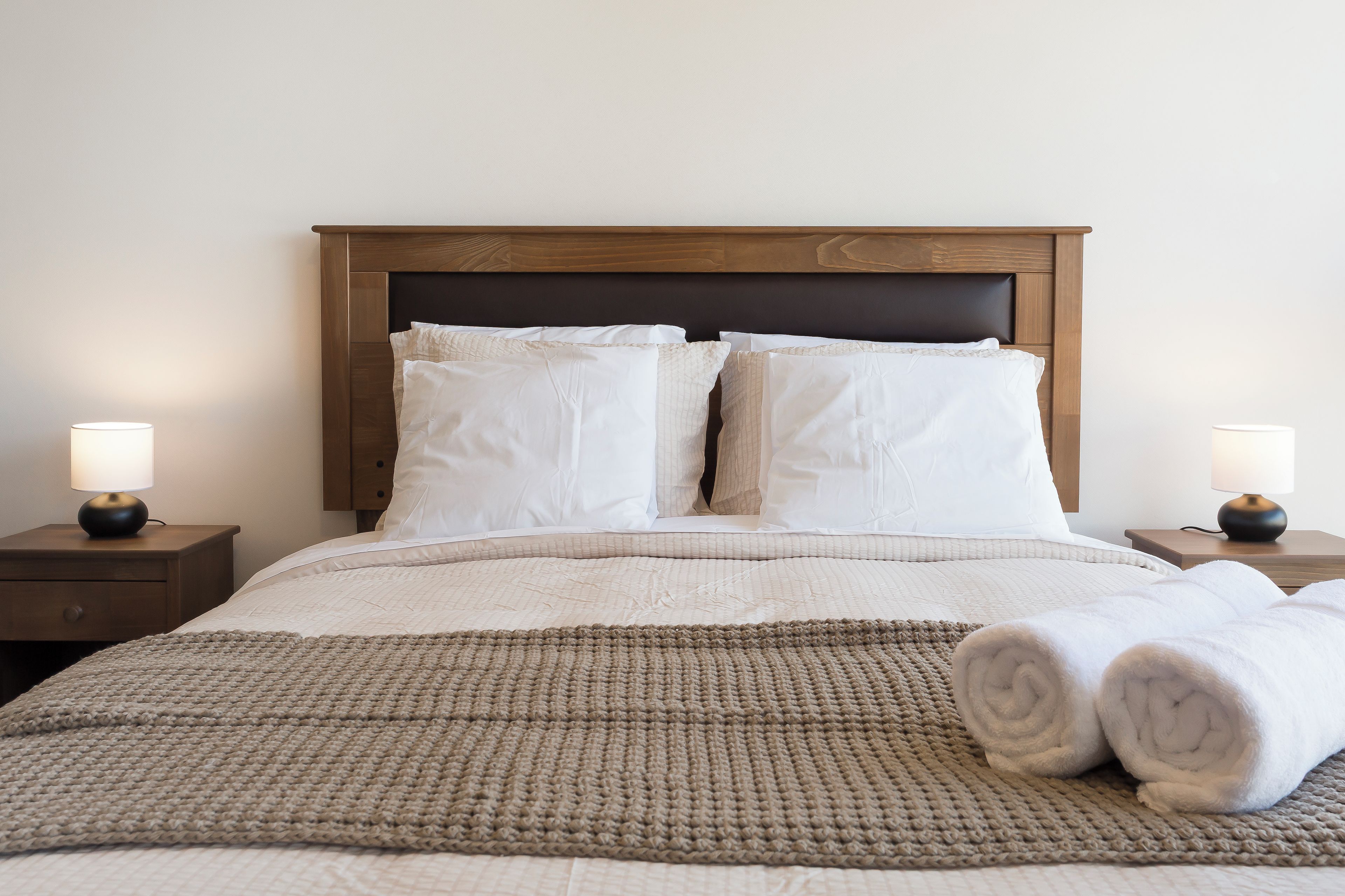 Una cama bien hecha con cabecera, dos lámparas de noche y una toalla enrollada sobre la cama.