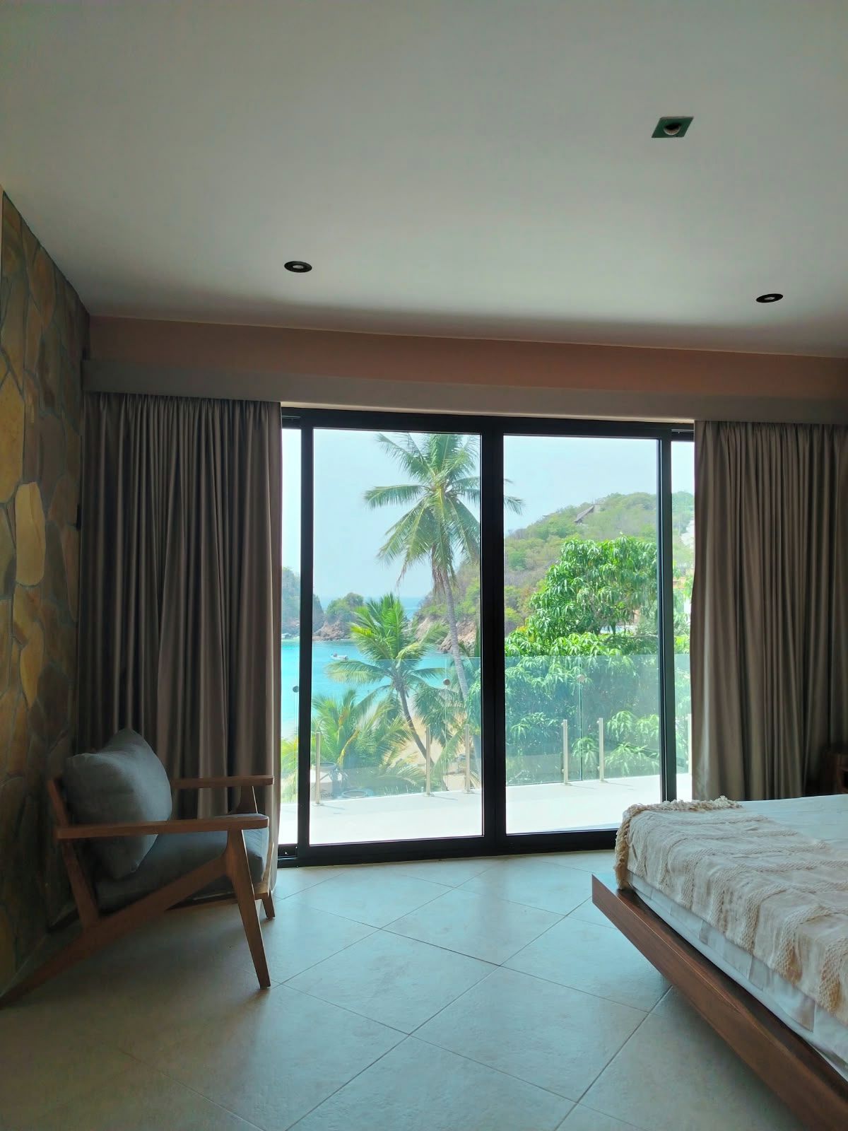 A bedroom with a large sliding glass door leading to a balcony with a view of a beach and palm trees.