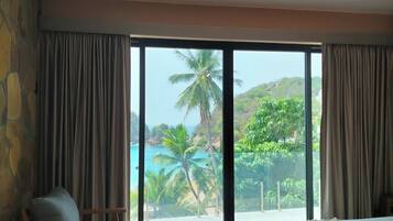 A bedroom with a large sliding glass door leading to a balcony with a view of a beach and palm trees.