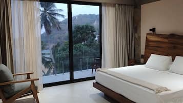 Quarto com cama grande, uma cadeira e vista para paisagem tropical através de janelas do chão ao teto.