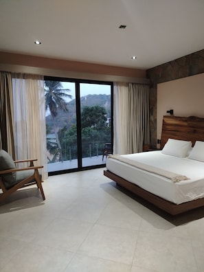 Un dormitorio con una cama grande, una silla y vistas a un paisaje tropical a través de ventanales de pared a techo.