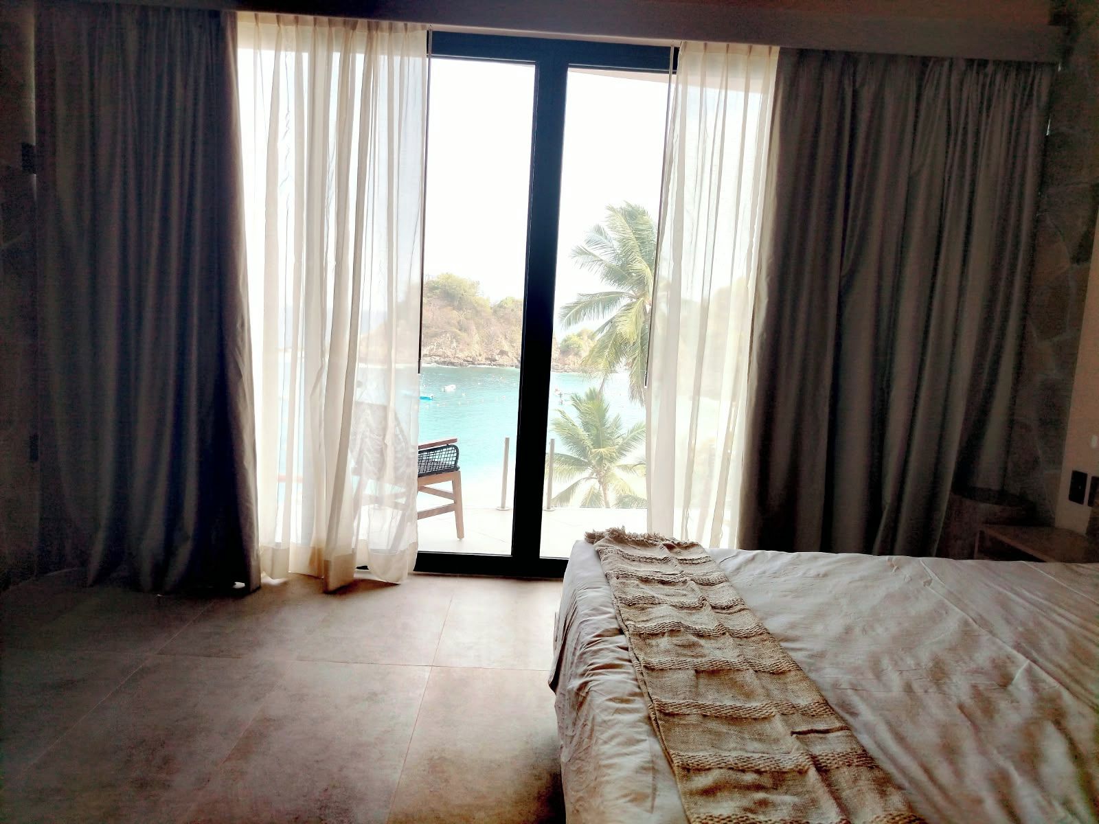 Un dormitorio con cama, una silla y vista a una playa y palmeras a través de un ventanal grande.