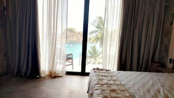 Un dormitorio con cama, una silla y vista a una playa y palmeras a través de un ventanal grande.