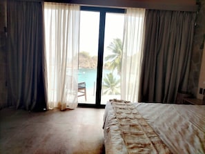 Un dormitorio con cama, una silla y vista a una playa y palmeras a través de un ventanal grande.
