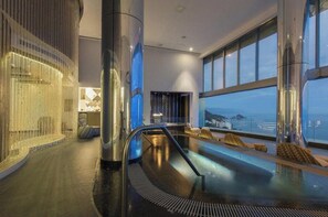 Un espace piscine intérieur moderne, doté d’une paroi en verre, de sièges et offrant une vue sur la mer.