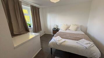 A bedroom with two beds, a window with curtains, a bedside table with a lamp, and a ceiling light fixture.