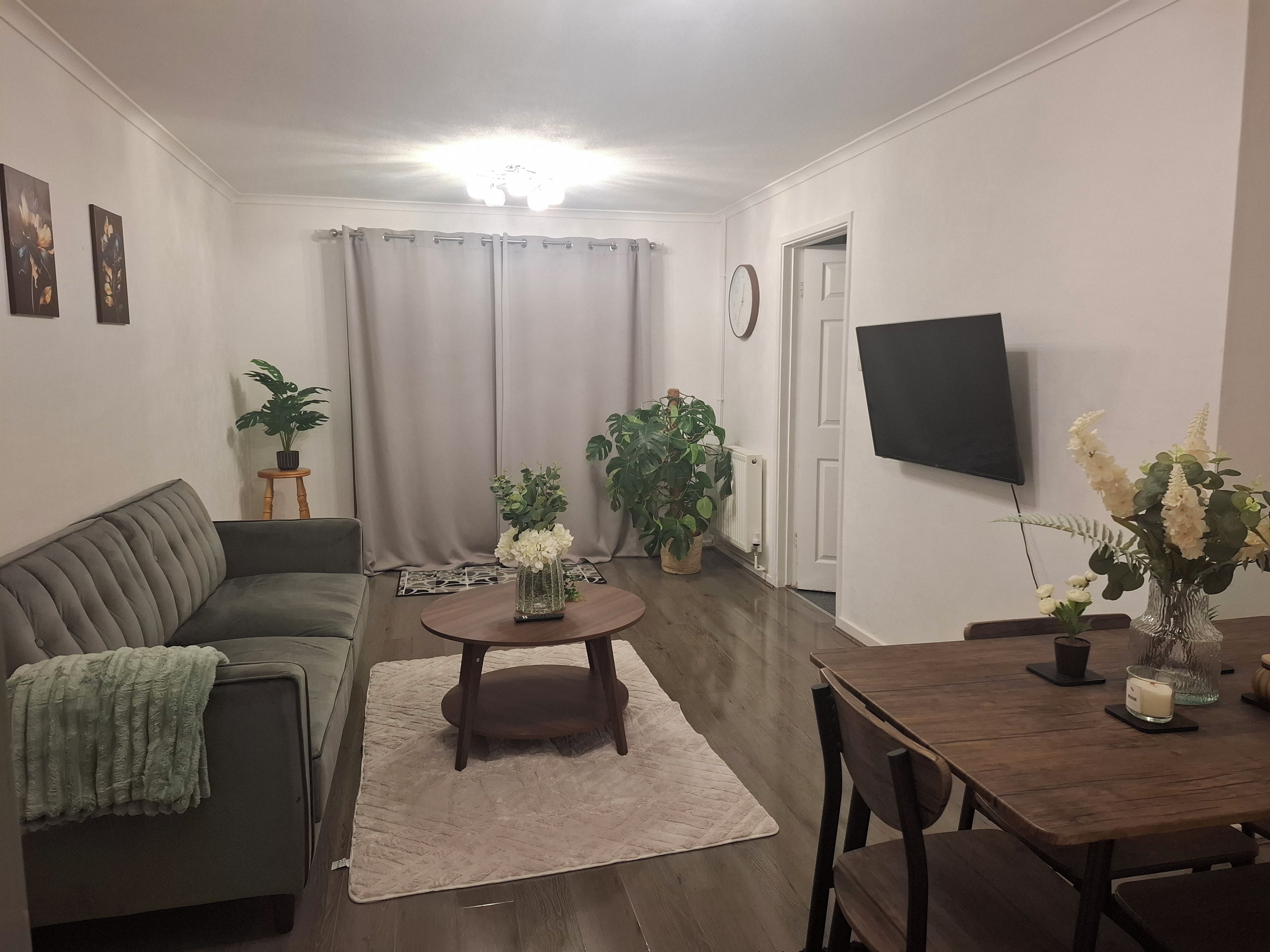 Ein Wohnzimmer mit einer Couch, einem Couchtisch, einem Esstisch mit Stühlen, einem an der Wand befestigten Fernseher und mehreren Kübelpflanzen.