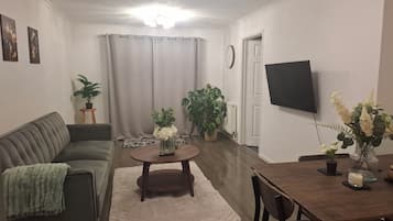 Ein Wohnzimmer mit einer Couch, einem Couchtisch, einem Esstisch mit Stühlen, einem an der Wand befestigten Fernseher und mehreren Kübelpflanzen.