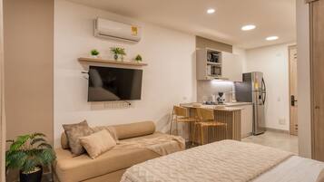 A modern living room with a sofa, a TV, a small kitchen area with a refrigerator, and a dining table.