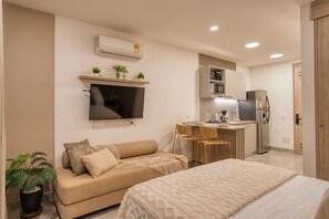 A modern living room with a sofa, a TV, a small kitchen area with a refrigerator, and a dining table.