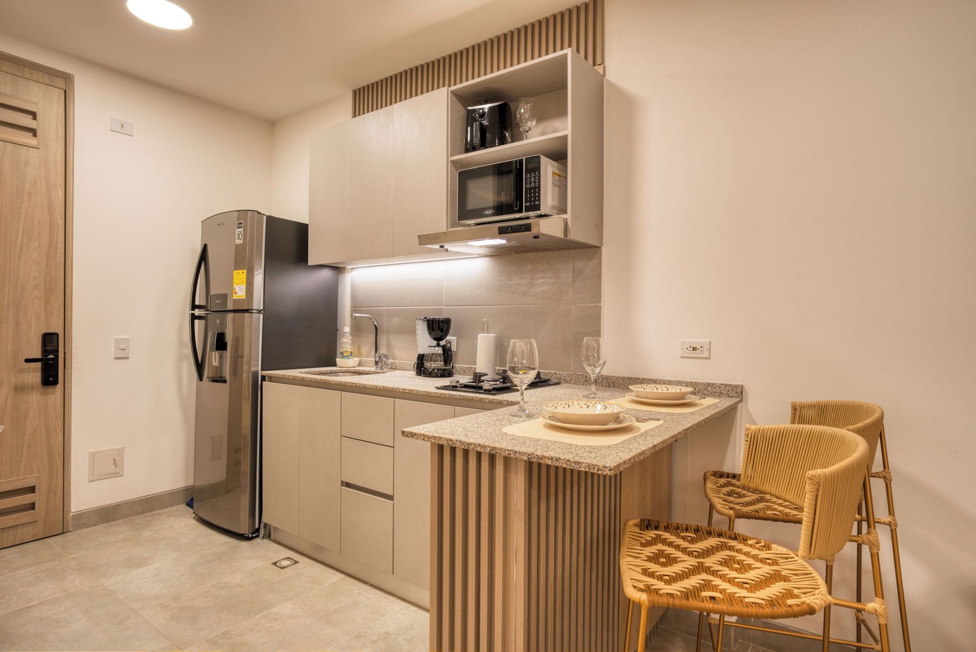 Cocina compacta con refrigerador integrado, microondas y cafetera. Cuenta con una pequeña barra con taburetes y una copa de vino.