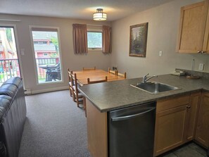 A compact kitchen with a built-in dishwasher, a dining area with a table and chairs, and a view through a glass door to the outside. - Alpine Escape | Ski-In/Out | Toby Chair | Pools (Panorama)