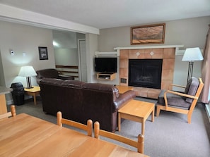 A living room with a brown leather sofa, a wooden coffee table, a wooden chair, a fireplace, and a television. - Alpine Escape | Ski-In/Out | Toby Chair | Pools (Panorama)