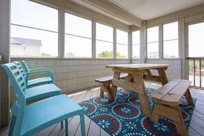 Eine überdachte Terrasse mit einem Holztisch und Stühlen, einem farbenfrohen Teppich und großen Fenstern.