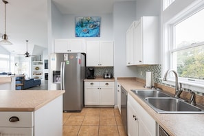 A modern kitchen with white cabinets, stainless steel appliances, a coffee maker, and a window with a view. - 2min Walk to Beach, Pool, Spa, Game Room, Big Yard (Corolla)