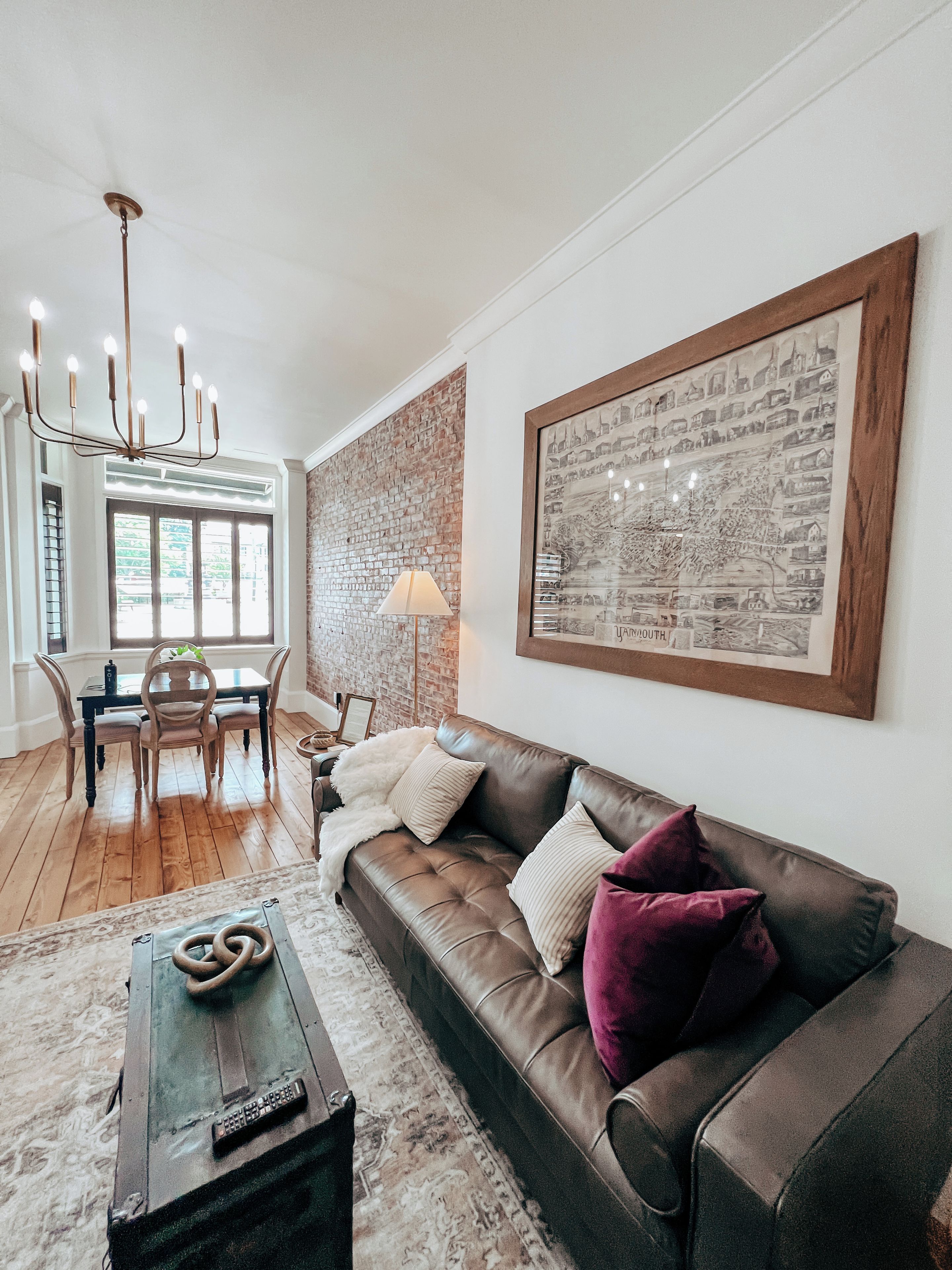 A living room with a brown leather sofa, a chandelier, a dining table with chairs, and a large framed picture on the wall.