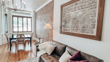 A living room with a brown leather sofa, a chandelier, a dining table with chairs, and a large framed picture on the wall.