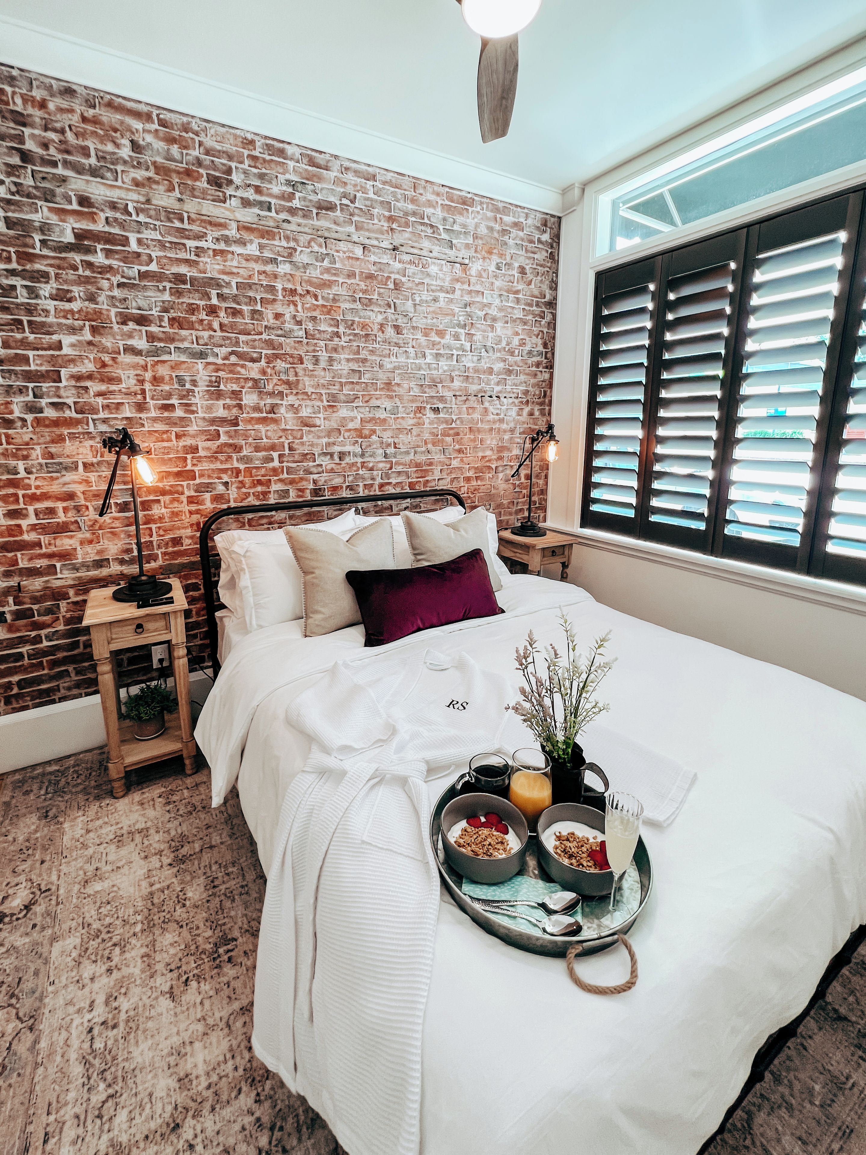 A bedroom with a brick wall, a bed with white bedding, a tray with breakfast items, and a nightstand with a lamp.