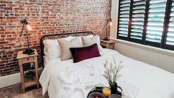 A bedroom with a brick wall, a bed with white bedding, a tray with breakfast items, and a nightstand with a lamp.