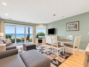 A living room with a sofa set, a dining table with chairs, a TV, and a view of the sea through large windows. - Bay Breeze 107 | Beachfront Condo | Near Downtown & Tart Trail (Traverse City)