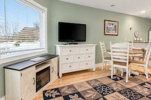 A room with a fireplace, a television, a dining table with chairs, and a patterned rug. - Bay Breeze 107 | Beachfront Condo | Near Downtown & Tart Trail (Traverse City)