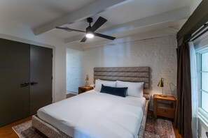 Ein Schlafzimmer mit einem Bett, einem Deckenventilator, einem Nachttisch, einer Lampe, einem Fenster mit Vorhängen und einer Ziegelwand.