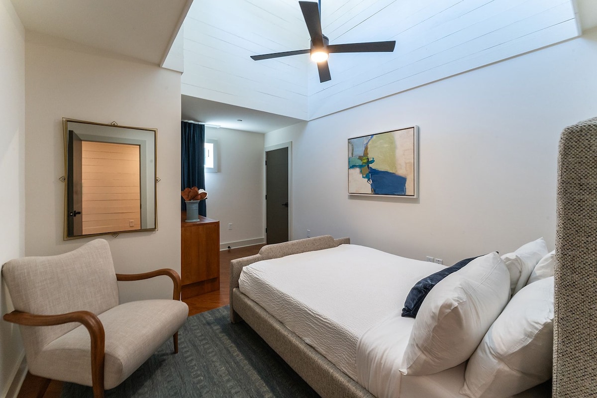 Ein Hotelzimmer mit Bett, Stuhl, Schreibtisch, Deckenventilator und einem Gemälde an der Wand.