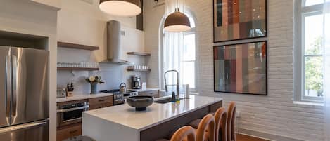 Una cocina moderna con una isla central, electrodomésticos de acero inoxidable, iluminación colgante y un ventanal grande.