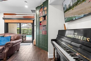 Ein Wohnzimmer mit einem Klavier, einem Ledersofa, einem Bücherregal und Blick auf den Strand durch ein Fenster.