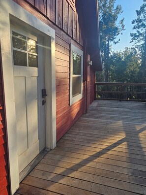 Una terraza de madera con una puerta blanca y una ventana, rodeada de árboles.