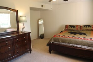 Une chambre à coucher avec une commode en bois, un lit recouvert d’une couette à motifs d’ours, un miroir et un ventilateur de plafond.