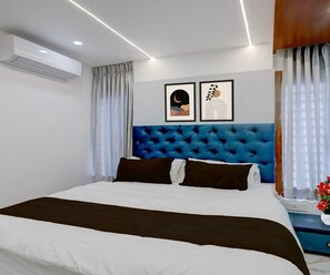 Ein modernes Hotelzimmer mit einem großen Bett, einem gepolsterten blauen Kopfteil, zwei gerahmten Kunstwerken und einem mit Vorhängen drapierten Fenster.