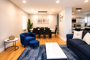 A modern living room with a blue sofa, a dining area with a table and chairs, and a kitchen in the background. - 8-Beds Big Stay, 10Mins NYC, Free Reserved Parking (Jersey City)