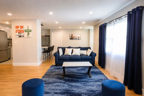 A modern living room with a sofa, coffee table, and ottomans, a kitchen in the background, and a dining area with chairs. - 8-Beds Big Stay, 10Mins NYC, Free Reserved Parking (Jersey City)