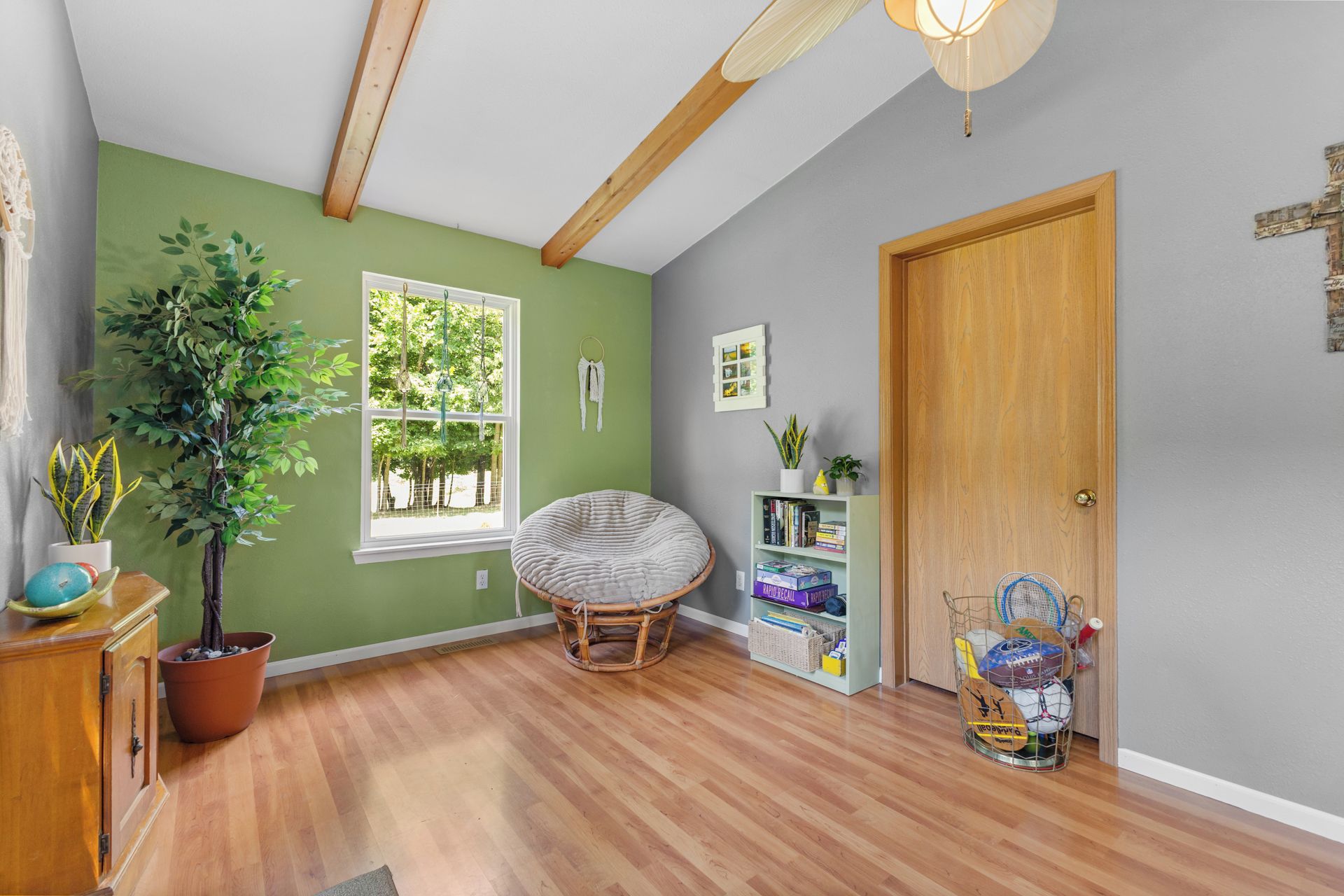 A room with a wooden floor, a green wall, a window, a door, and a bookshelf with books and a plant.
