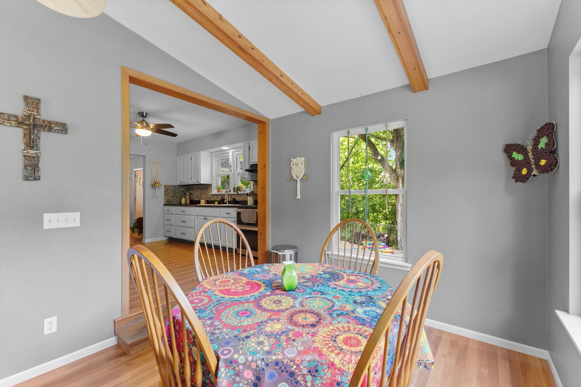 Un comedor con una mesa redonda, sillas y un mantel colorido. Se observa una ventana con vistas a árboles y una decoración con forma de mariposa en la pared.