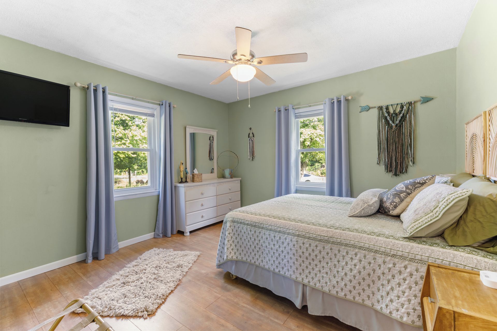 A bedroom with a bed, dresser, mirror, ceiling fan, windows, and a rug.